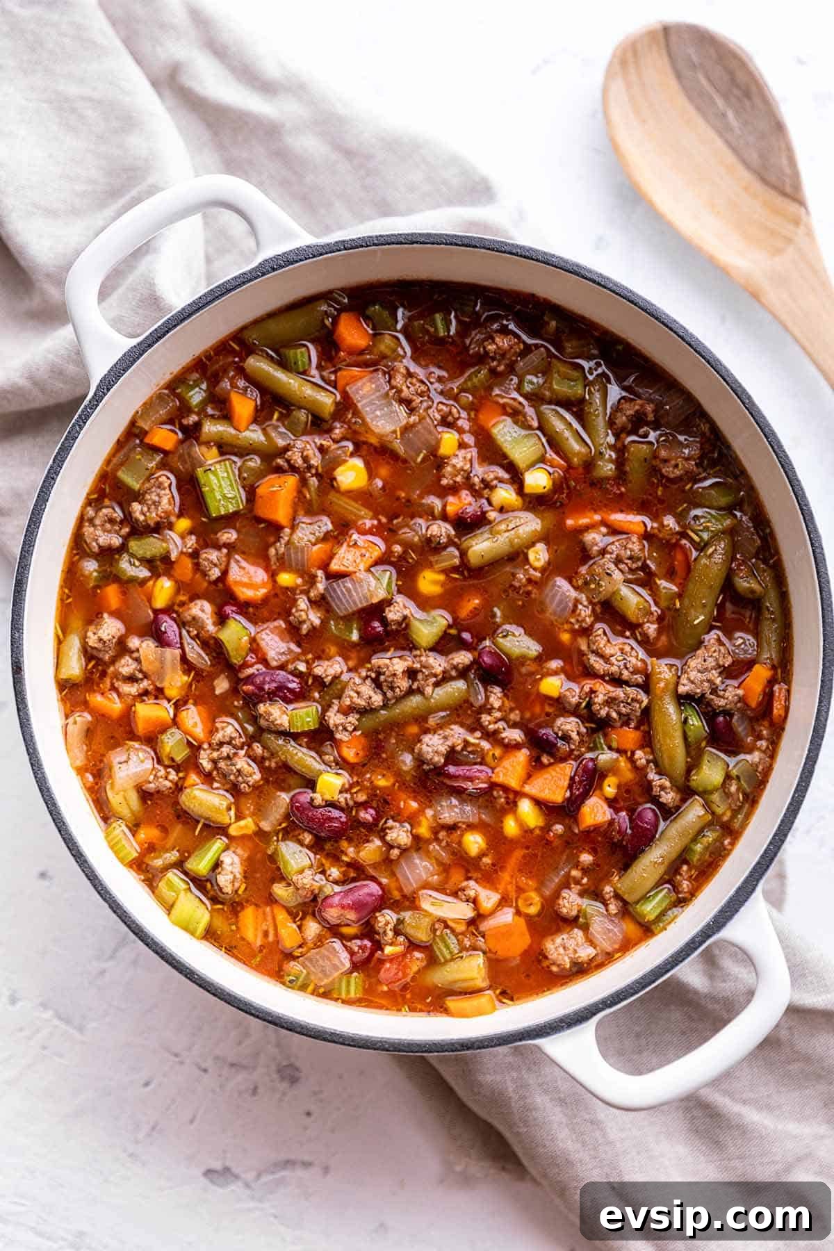A white Dutch Oven full of a homemade soup with a wooden spoon on the side.