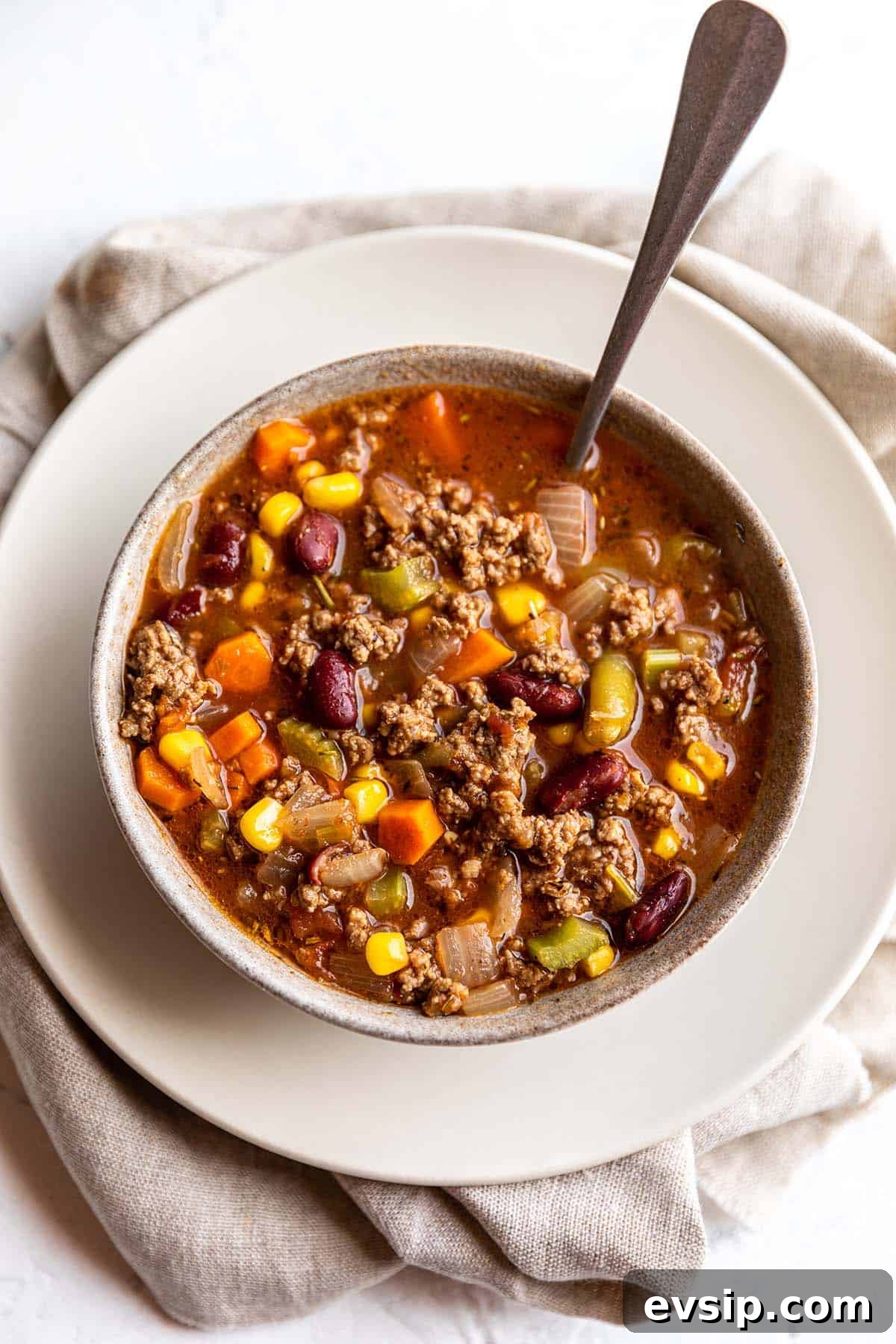 A bowl full of a soup that is green beans, corn, carrots, celery and ground beef.