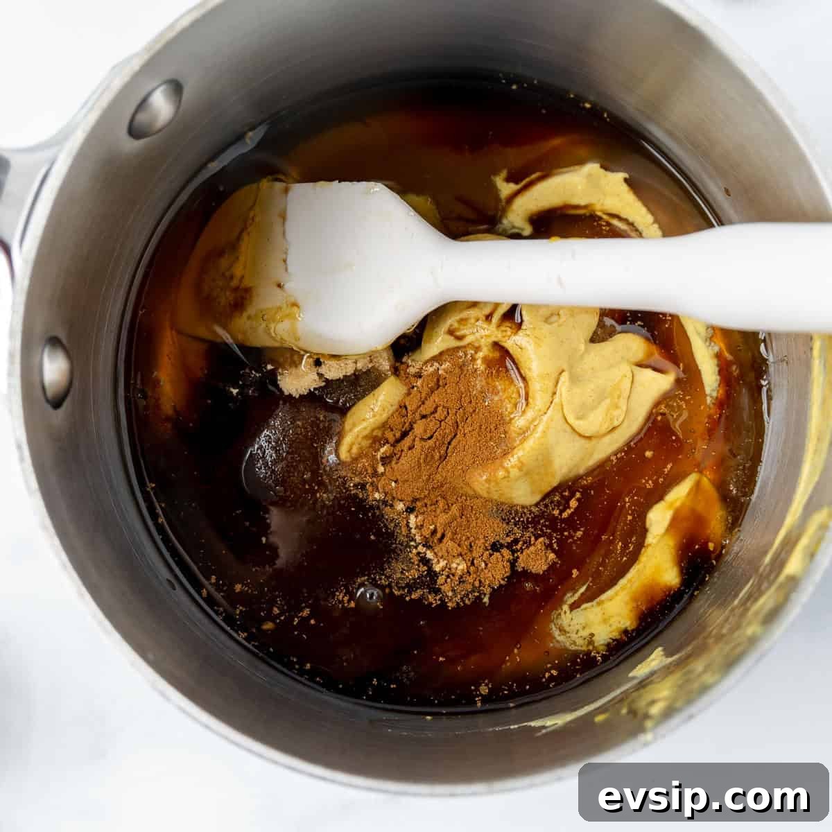 Ingredients including spices, mustard, and liquid blend in a saucepan with a white spatula.