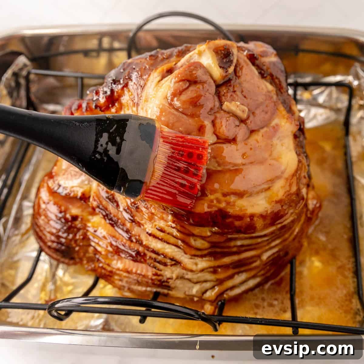 A baked ham is being glazed with a red brush on a rack over a pan.