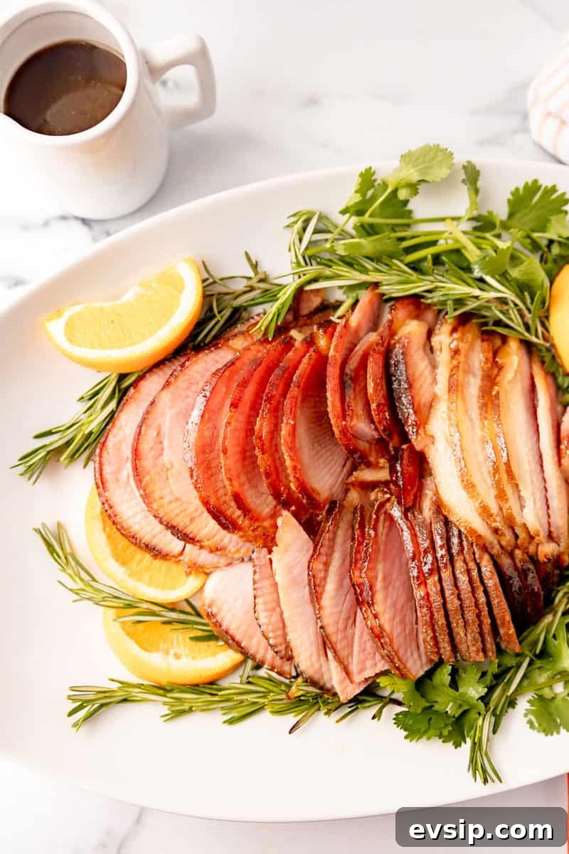 Sliced ham garnished with rosemary and orange slices on a white platter. A small cup of sauce is in the background.
