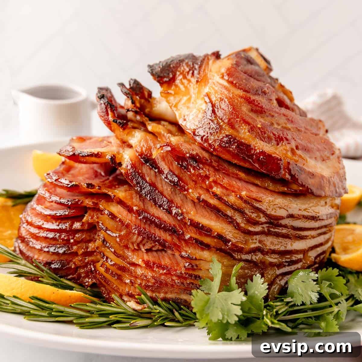 A glazed spiral ham garnished with herbs and orange slices on a white plate, with a sauce jug in the background.
