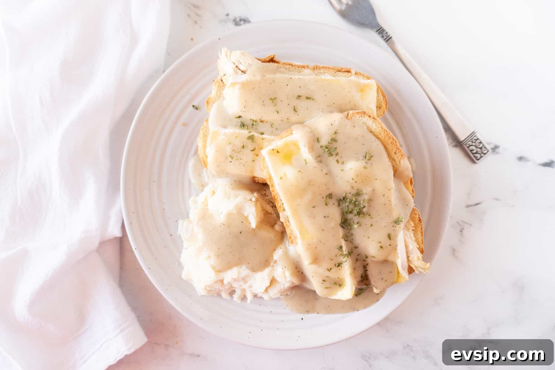 Hot open-faced turkey sandwich with gravy on a white plate next to mashed potatoes, a plate next to a fork, and a napkin.