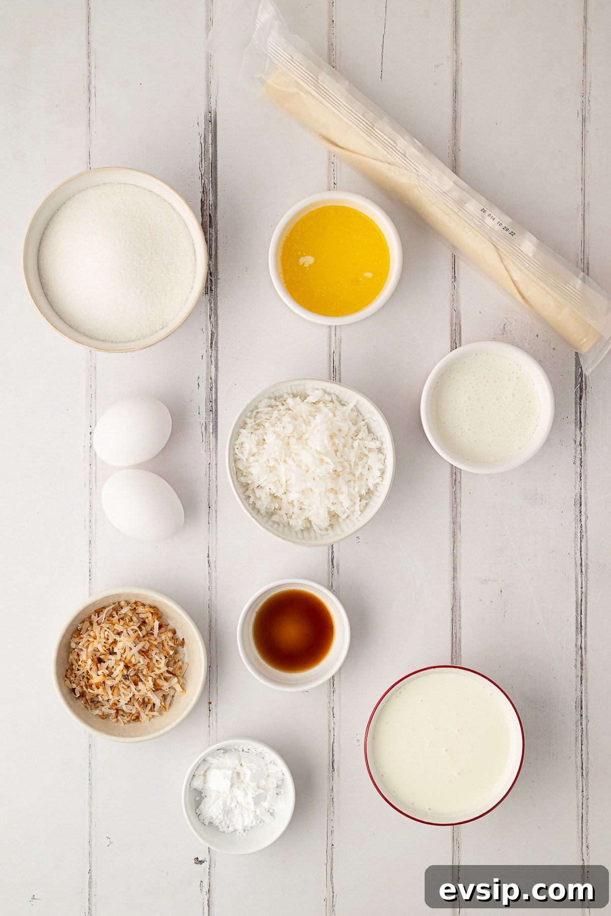Ingredients needed to make an easy coconut pie, including a bowl of shredded coconut, an unbaked pie crust, buttermilk, sugar, butter, and eggs arranged on a kitchen counter.