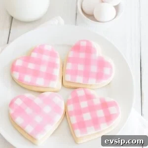 A plate of four heart-shaped cookies decorated with pink and white gingham icing. The plate is on a white cloth. In the background, there are eggs, a glass of milk, and a jar. The setting is on a white wooden surface.