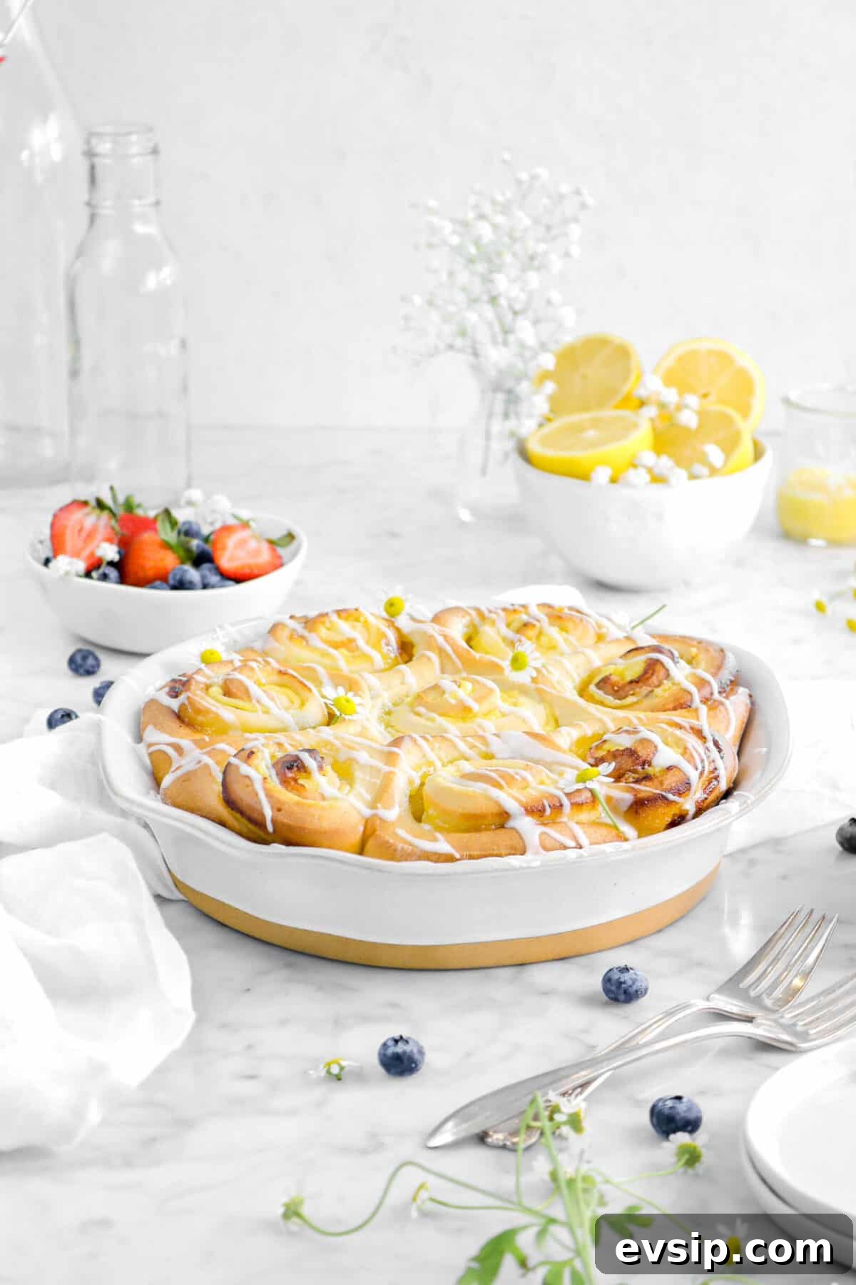 lemon sweet rolls in pie pan with berries, fruit, a bowl of lemons, flowers, and two glasses