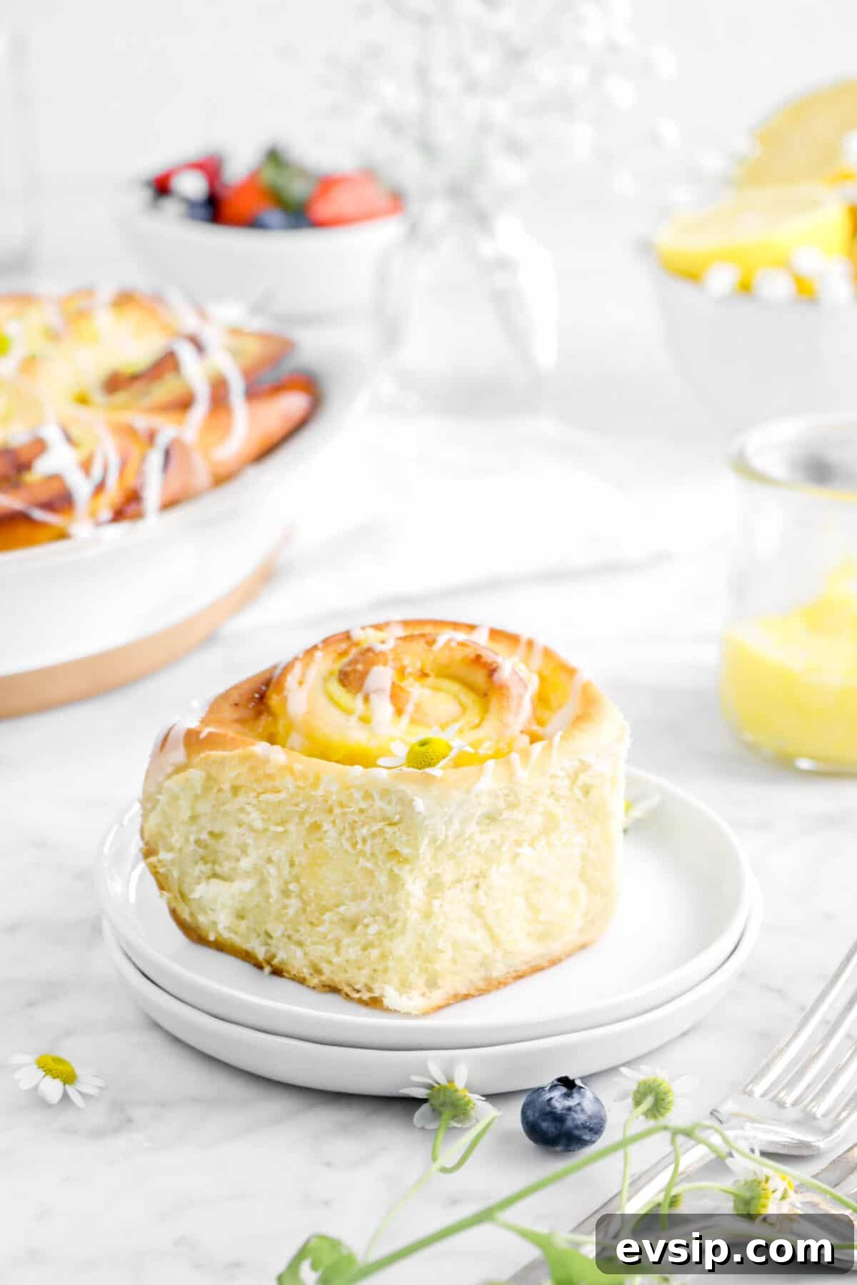lemon sweer roll on two plates with flowers, lemon custard, berries, and rolls behind