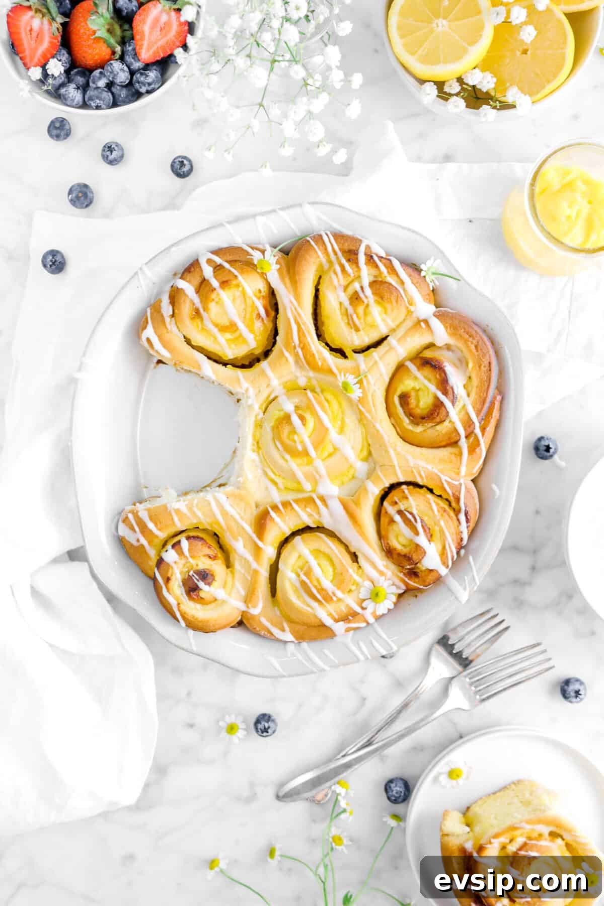 overhead shot of lemon sweet rolls with one taken out of the pan with berries, a white napkin, lemon curd, and forks