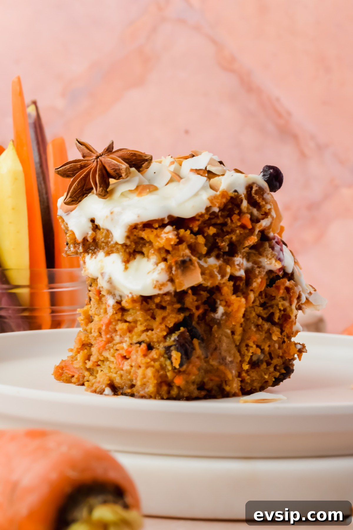 slice of two layer carrot cake on white plate with peeled carrots in background.