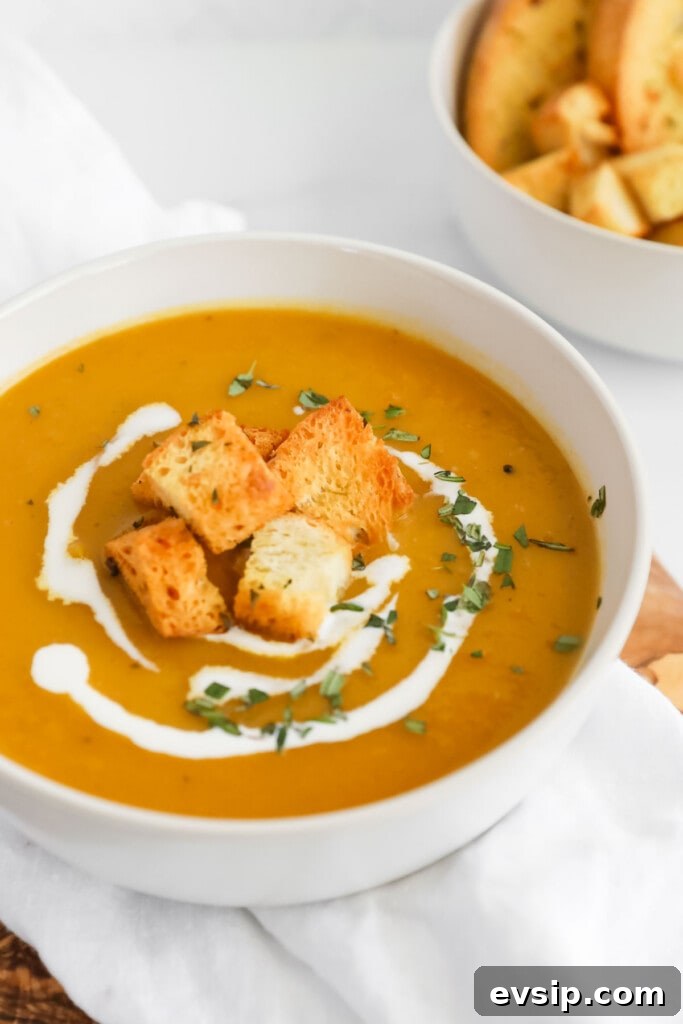 A close up image of a white bowl filled with orange soup. On top of the soup there are croutons, freshly chopped rosemary, and a drizzle of coconut milk.